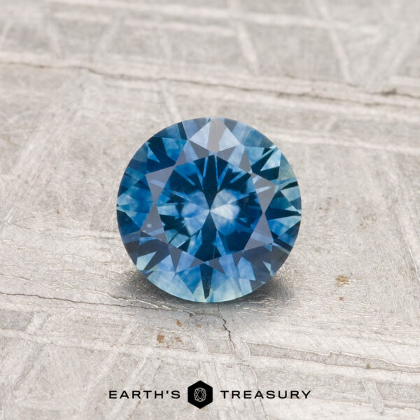0.88-Carat Montana Sapphire (Heated) 0.88-Carat Montana Sapphire (Heated), round brilliant-cut, blue gemstone displayed on a textured gray surface with "Earth’s Treasury" logo at the bottom.