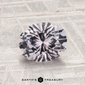 A 1.87-carat Montana sapphire with a round brilliant cut and faceted surface rests on a textured light gray background. The "Earth's Treasury" logo appears at the bottom of the image.