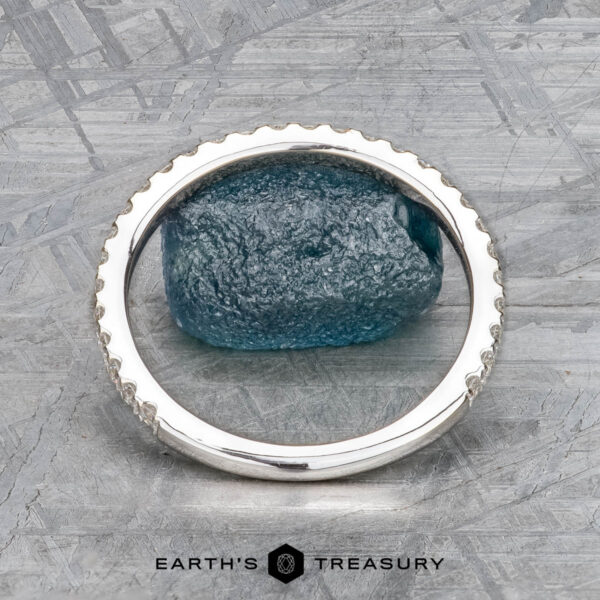 The "Amphitrite" band in 18K white gold A textured blue rough gemstone is set in the "Amphitrite" band, crafted in 18K white gold with a notched edge, displayed on a mottled gray surface. "Earth's Treasury" is shown at the bottom.