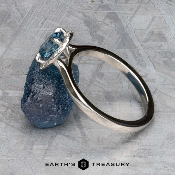 The "Alice" ring in platinum with 1.09-carat Montana sapphire The "Alice" ring in platinum with a 1.09-carat Montana sapphire is displayed against a rough blue stone on a textured gray surface. The Earth's Treasury logo appears at the bottom.