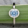 The "Alice" ring in platinum, featuring a 1.09-carat Montana sapphire surrounded by a diamond halo, is displayed on a green surface.