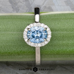 The "Alice" ring in platinum, featuring a 1.09-carat Montana sapphire surrounded by a diamond halo, is displayed on a green surface.