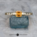 The "Karelia" ring in 14K yellow gold features a 0.52-carat rectangular Montana sapphire, accented with six small diamonds, displayed on a rough blue stone atop a textured gray surface.