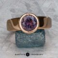 The "Declan" ring in 20K pink gold, featuring a 2.81-carat Montana sapphire, is shown resting on a blue textured stone against a metallic patterned background.