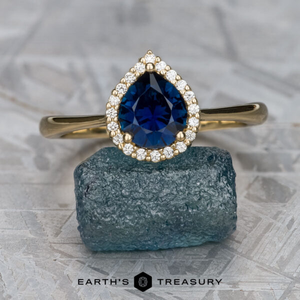 The "Margaret" ring in 14K yellow gold with 1.06-Carat Nigerian Sapphire The "Margaret" ring in 14K yellow gold with a 1.06-carat Nigerian sapphire, surrounded by small diamonds, is displayed on a rough blue gemstone, with the text "EARTH'S TREASURY" below.