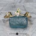 The "Jasmine" band in 14K yellow gold, adorned with green and clear gemstones, rests on a rough blue stone against a textured grey background. "EARTH'S TREASURY" is featured at the bottom.