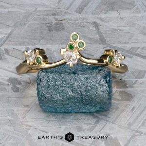 The "Jasmine" band in 14K yellow gold, adorned with green and clear gemstones, rests on a rough blue stone against a textured grey background. "EARTH'S TREASURY" is featured at the bottom.