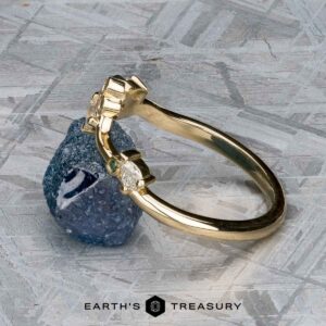 The "Jasmine" band in 14K yellow gold, set with a clear gemstone, is displayed upright against a rough blue stone on a textured gray surface. "EARTH'S TREASURY" appears at the bottom.