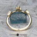 The “Jasmine” band in 14K yellow gold stands upright before a rough blue gemstone on a textured gray surface, with the "EARTH'S TREASURY" logo displayed at the bottom.