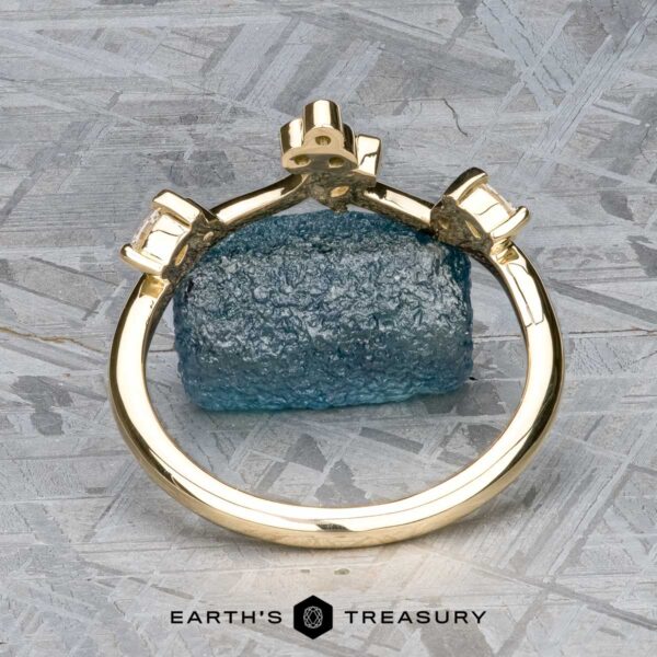 The "Jasmine" band in 14K yellow gold The “Jasmine” band in 14K yellow gold stands upright before a rough blue gemstone on a textured gray surface, with the "EARTH'S TREASURY" logo displayed at the bottom.