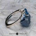 The "Lamia" ring in platinum with a 1.36-carat Montana sapphire is shown leaning against a rough blue mineral on a textured surface, with two smaller side stones and the Earth's Treasury logo at the bottom.