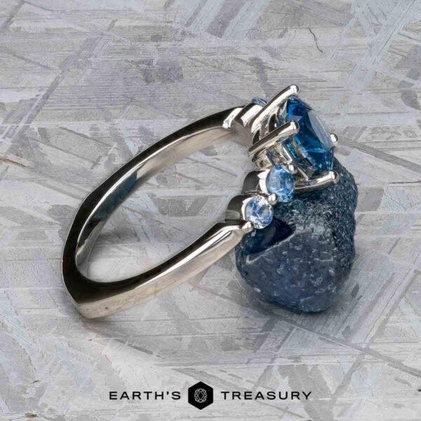 The "Lamia" ring in platinum with 1.36-carat Montana sapphire The "Lamia" ring in platinum with a 1.36-carat Montana sapphire is shown leaning against a rough blue mineral on a textured surface, with two smaller side stones and the Earth's Treasury logo at the bottom.