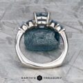 The "Lamia" ring in platinum with a 1.36-carat Montana sapphire stands upright before a rough blue stone on a textured gray surface, with the Earth's Treasury logo below.
