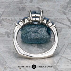 The "Lamia" ring in platinum with a 1.36-carat Montana sapphire stands upright before a rough blue stone on a textured gray surface, with the Earth's Treasury logo below.