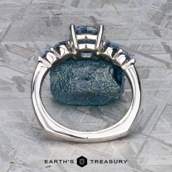 The "Lamia" ring in platinum with 1.36-carat Montana sapphire The "Lamia" ring in platinum with a 1.36-carat Montana sapphire stands upright before a rough blue stone on a textured gray surface, with the Earth's Treasury logo below.