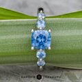 The "Lamia" ring in platinum, featuring a 1.36-carat cushion-cut Montana sapphire and four round blue stones, is displayed on a green leaf. The "EARTH'S TREASURY" logo appears at the bottom.