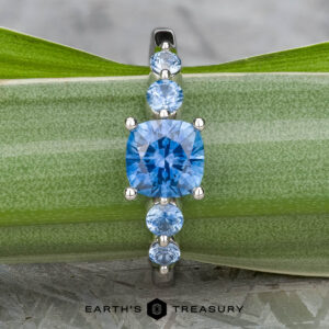 The "Lamia" ring in platinum, featuring a 1.36-carat cushion-cut Montana sapphire and four round blue stones, is displayed on a green leaf. The "EARTH'S TREASURY" logo appears at the bottom.