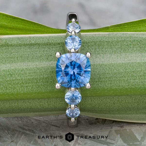 The "Lamia" ring in platinum with 1.36-carat Montana sapphire The "Lamia" ring in platinum, featuring a 1.36-carat cushion-cut Montana sapphire and four round blue stones, is displayed on a green leaf. The "EARTH'S TREASURY" logo appears at the bottom.