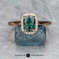 The "Alice" ring in 14K rose gold, featuring a 1.50-carat Australian sapphire and diamond halo, stands upright on a rough blue stone with a textured gray background. The "Earth's Treasury" logo and text are shown below.