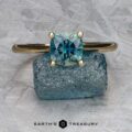 The "Katya" ring in 14K yellow gold, featuring a 1.35-carat Australian sapphire, rests on a rough blue stone against a textured gray background. The "Earth's Treasury" logo appears at the bottom.