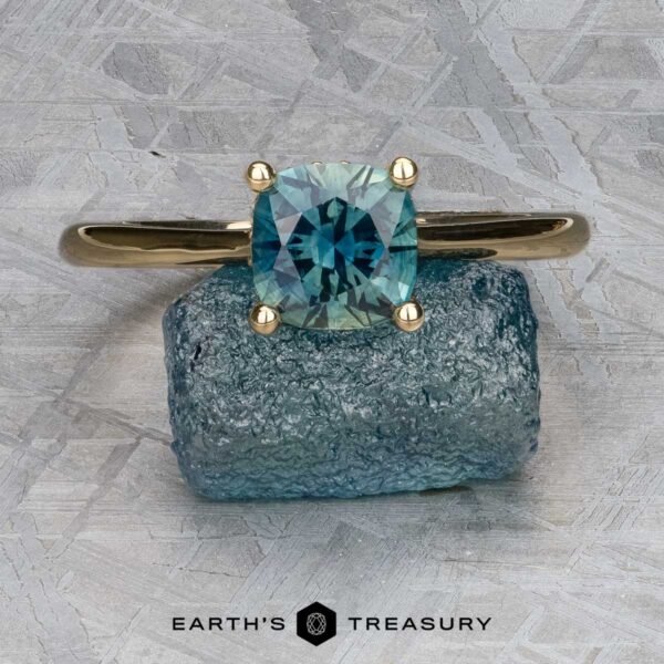 The "Katya" ring in 14K yellow gold with 1.35-carat Australian sapphire The "Katya" ring in 14K yellow gold, featuring a 1.35-carat Australian sapphire, rests on a rough blue stone against a textured gray background. The "Earth's Treasury" logo appears at the bottom.