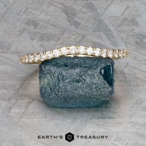 The Petite Pave Contour Band in 14K yellow gold is displayed on a rough blue gemstone against a textured gray background. The text "EARTH'S TREASURY" appears at the bottom.