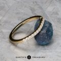 The Petite Pave Contour Band in 14K yellow gold, featuring small diamonds, rests against a rough blue gemstone on a textured gray surface, with the "EARTH'S TREASURY" logo displayed below.