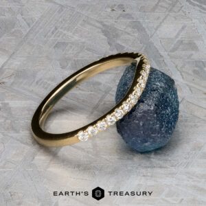 The Petite Pave Contour Band in 14K yellow gold, featuring small diamonds, rests against a rough blue gemstone on a textured gray surface, with the "EARTH'S TREASURY" logo displayed below.