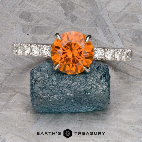 The Deluxe Pave "Claire" ring in platinum with 2.05-carat Montana sapphire A platinum Deluxe Pave "Claire" ring, set with a 2.05-carat Montana sapphire center and small clear stones on the band, is displayed on a rough blue mineral. Text below reads "Earth's Treasury.
