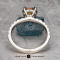 The Deluxe Pave "Claire" ring in platinum, featuring a 2.05-carat Montana sapphire, is displayed upright against a rough blue-green stone, with the "Earth's Treasury" logo at the bottom.
