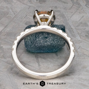 The Deluxe Pave "Claire" ring in platinum, featuring a 2.05-carat Montana sapphire, is displayed upright against a rough blue-green stone, with the "Earth's Treasury" logo at the bottom.