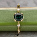 The "Pirouette" ring features a 1.17-carat round Australian Sapphire set in 18K yellow gold, accented by small white gems along the band, and is shown displayed on a green surface.