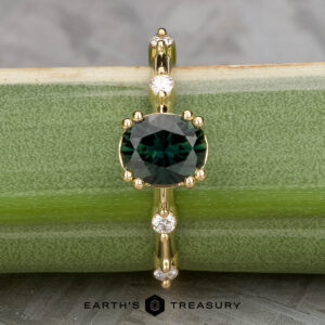 The "Pirouette" ring features a 1.17-carat round Australian Sapphire set in 18K yellow gold, accented by small white gems along the band, and is shown displayed on a green surface.
