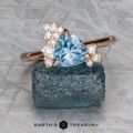 The "Praesepe" ring in 14K rose gold, featuring a 1.29-carat triangular Montana sapphire and small round diamonds, is displayed on a rough blue stone slab against a textured gray background.