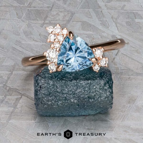 The "Praesepe" ring in 14K rose gold with 1.29-carat Montana sapphire The "Praesepe" ring in 14K rose gold, featuring a 1.29-carat triangular Montana sapphire and small round diamonds, is displayed on a rough blue stone slab against a textured gray background.