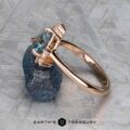 The "Praesepe" ring in 14K rose gold, featuring a 1.29-carat Montana sapphire and small diamonds, rests on a rough dark mineral atop a textured surface with the Earth's Treasury logo below.