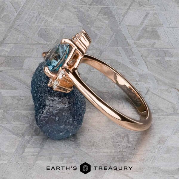 The "Praesepe" ring in 14K rose gold with 1.29-carat Montana sapphire The "Praesepe" ring in 14K rose gold, featuring a 1.29-carat Montana sapphire and small diamonds, rests on a rough dark mineral atop a textured surface with the Earth's Treasury logo below.