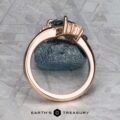The "Praesepe" ring in 14K rose gold, featuring a 1.29-carat Montana sapphire and baguette side stones, stands upright on a textured gray surface. The "Earth's Treasury" logo is visible at the bottom.