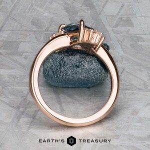 The "Praesepe" ring in 14K rose gold, featuring a 1.29-carat Montana sapphire and baguette side stones, stands upright on a textured gray surface. The "Earth's Treasury" logo is visible at the bottom.