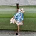 The "Praesepe" ring—a 1.29-carat Montana sapphire set in 14K rose gold—is shown against green bamboo. A cluster of round white diamonds sparkles beside the triangular blue gemstone, with the "Earth's Treasury" logo beneath.