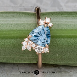 The "Praesepe" ring—a 1.29-carat Montana sapphire set in 14K rose gold—is shown against green bamboo. A cluster of round white diamonds sparkles beside the triangular blue gemstone, with the "Earth's Treasury" logo beneath.