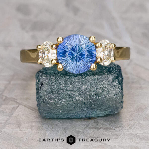 The "Evelyn" ring in 18K yellow gold with 1.27-carat Ceylon sapphire The "Evelyn" ring in 18K yellow gold features a 1.27-carat Ceylon sapphire center and two oval clear side stones, displayed on a rough blue stone. "Earth's Treasury" text is shown at the bottom.