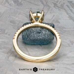 The Tapered Pave "Claire" ring in 18k yellow gold with a 1.36-carat Montana sapphire stands upright on a blue-grey stone, viewed from the side. The "Earth's Treasury" logo appears at the bottom.