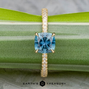 The Tapered Pave "Claire" ring in 18k yellow gold features a 1.36-carat Montana sapphire and diamond accents on the band, displayed on a green plant. Logo text: "Earth's Treasury.