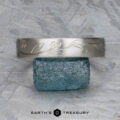 The "Olive" band in platinum, featuring an engraved leaf pattern, rests on a rough blue gemstone. The background is a textured gray surface. "EARTH'S TREASURY" appears below the objects.