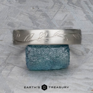 The "Olive" band in platinum, featuring an engraved leaf pattern, rests on a rough blue gemstone. The background is a textured gray surface. "EARTH'S TREASURY" appears below the objects.