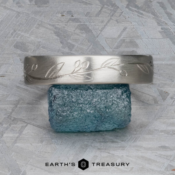 The "Olive" band in platinum The "Olive" band in platinum, featuring an engraved leaf pattern, rests on a rough blue gemstone. The background is a textured gray surface. "EARTH'S TREASURY" appears below the objects.