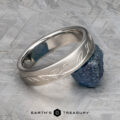 The "Olive" band in platinum, featuring an engraved leaf pattern, rests against a raw blue gemstone on a textured gray surface. "EARTH'S TREASURY" appears at the bottom.