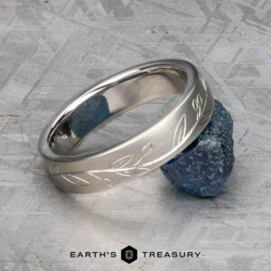 The "Olive" band in platinum, featuring an engraved leaf pattern, rests against a raw blue gemstone on a textured gray surface. "EARTH'S TREASURY" appears at the bottom.
