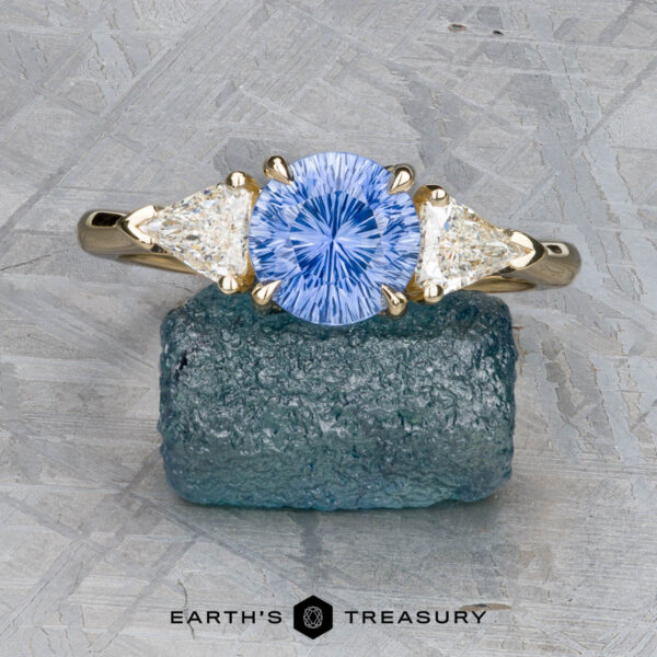 The “Mielikki” ring in 14K yellow gold with 1.42-Carat Ceylon sapphire The “Mielikki” ring in 14K yellow gold features a 1.42-carat Ceylon sapphire flanked by two triangle-cut clear stones, displayed on a raw mineral with a geometric background.